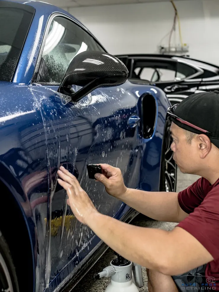 A man is applying a protective film to the side of a blue sports car.