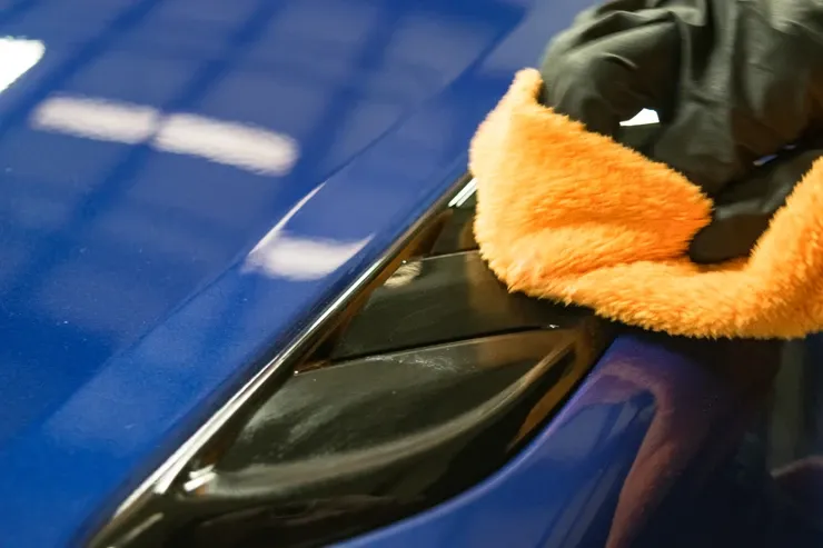 A person wearing black gloves is cleaning a blue car with an orange towel