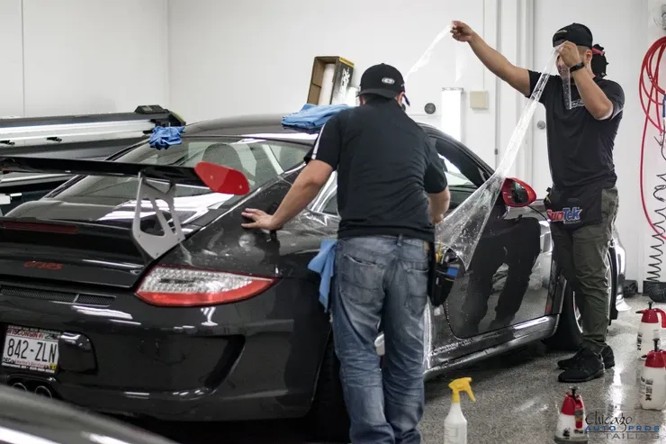 Two men are wrapping a black sports car in a garage.