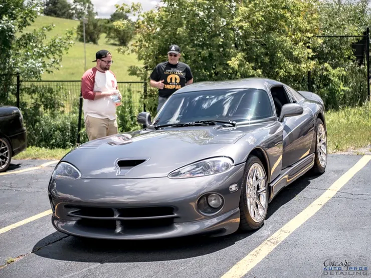 A gray dodge viper is parked in a parking lot with two men standing next to it.