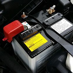 Car battery in engine compartment, with red terminal cover and black hold-down bracket.