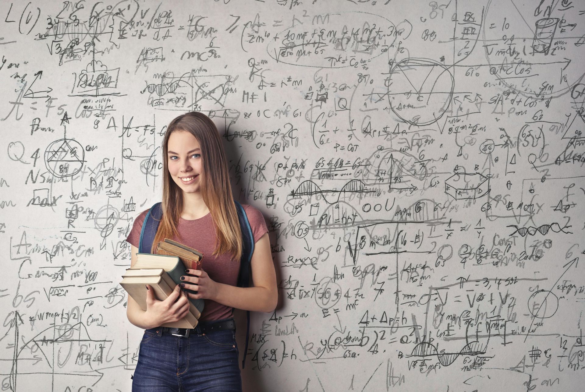 A white female holding university books is standing in front of a white board that showcases very complex mathematical equations. 