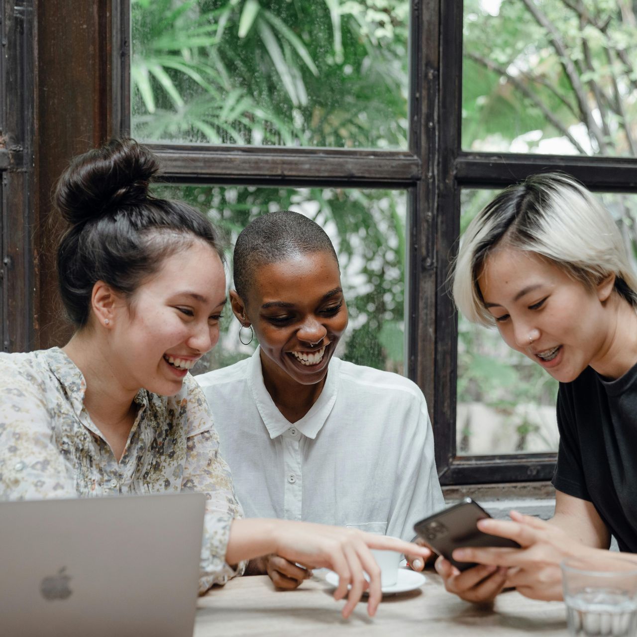 Three women are sitting at a table with a laptop and a cell phone.