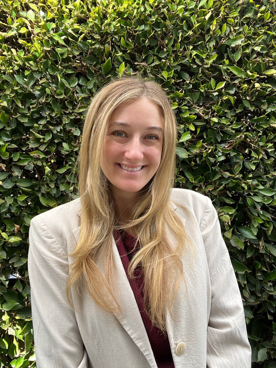 Woman with long blonde hair, smiling, wearing a cream blazer and maroon top, standing in front of a green hedge.