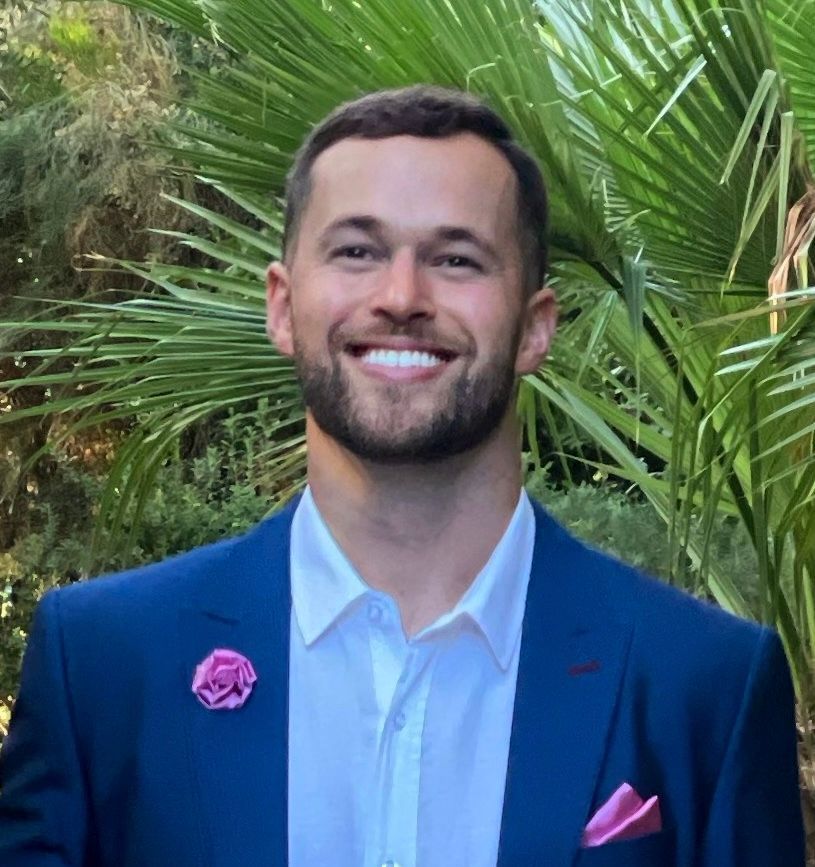 Man in blue suit, white shirt, and pink lapel flower smiles against a leafy background.