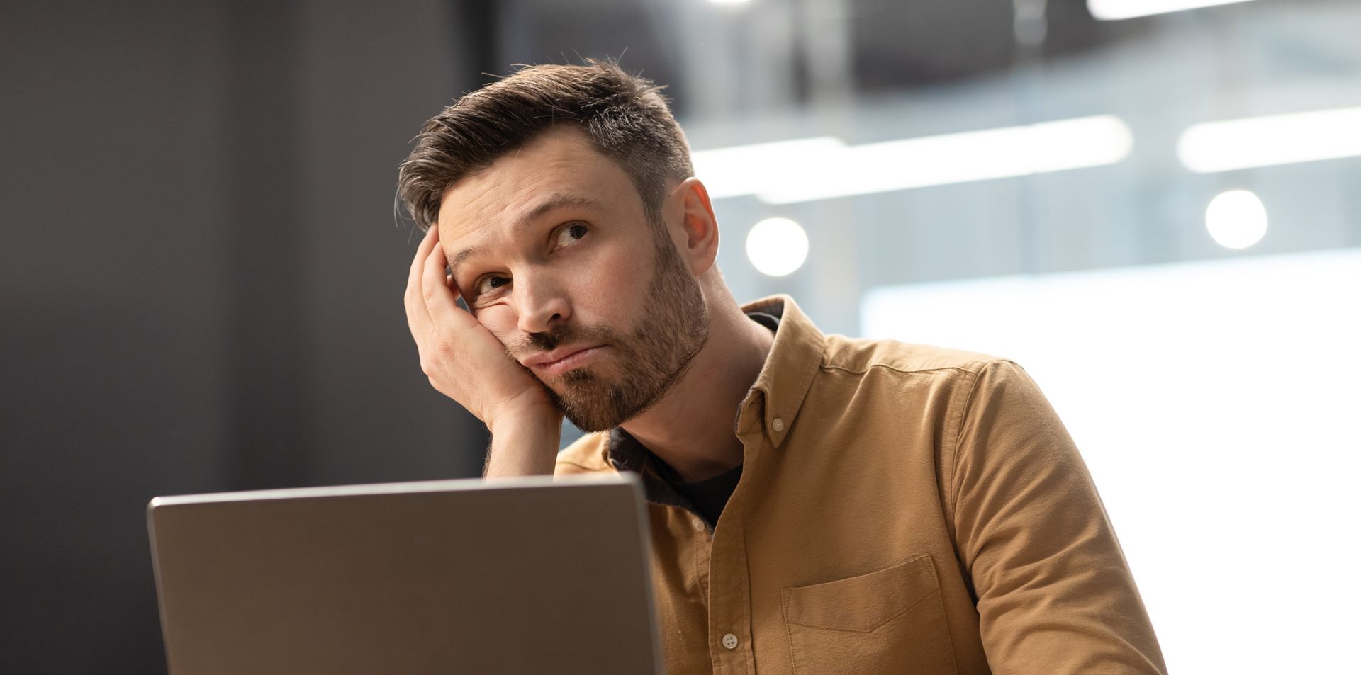A man is sitting in front of a laptop computer with his hand on his face.