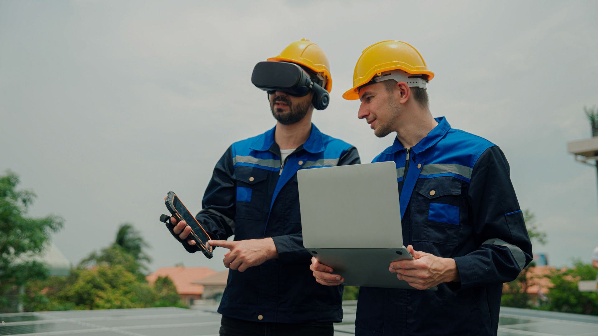 Two construction workers wearing hard hats and virtual reality glasses are looking at a laptop.