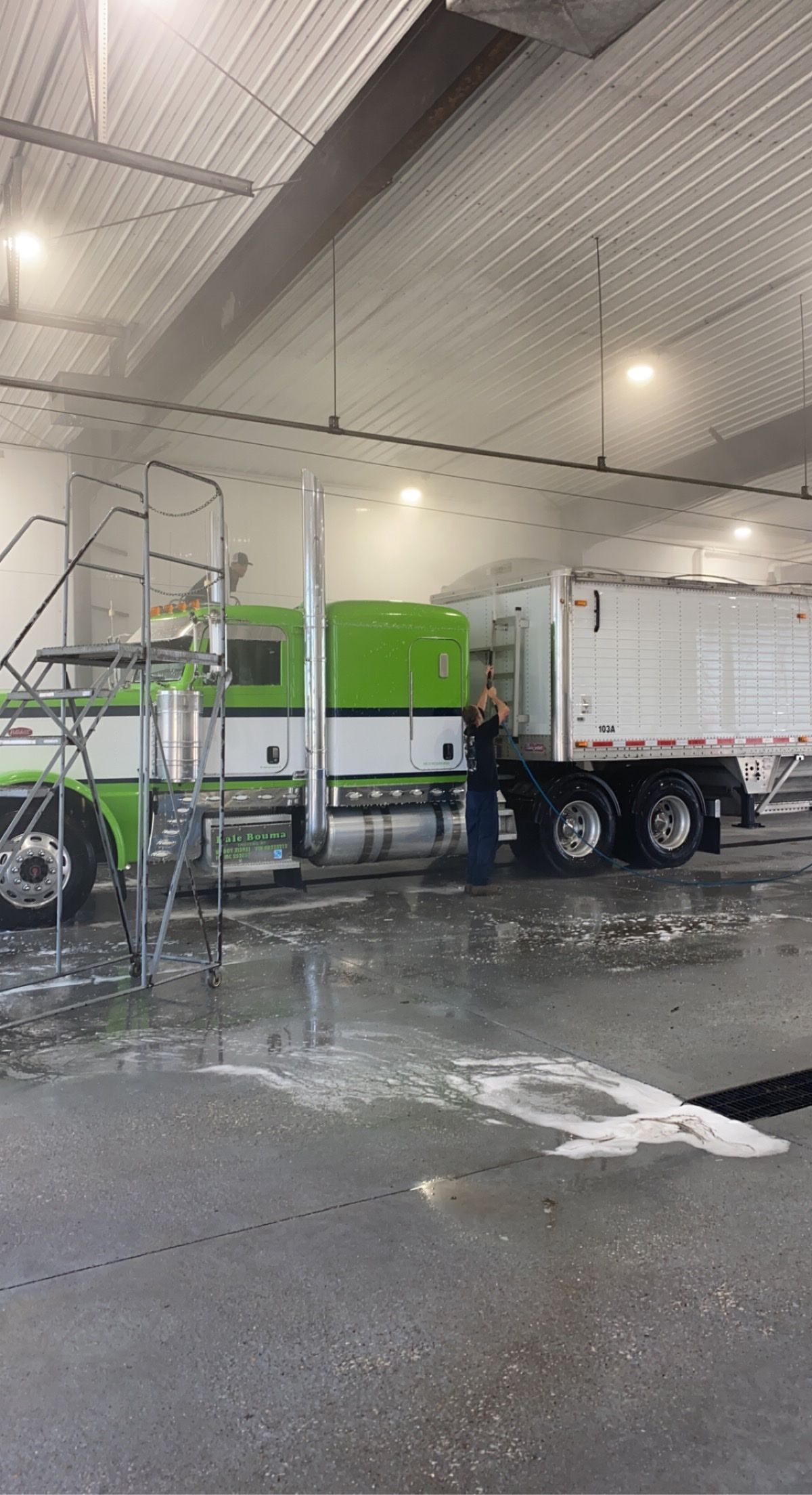 A green and white semi truck is being washed in a warehouse.