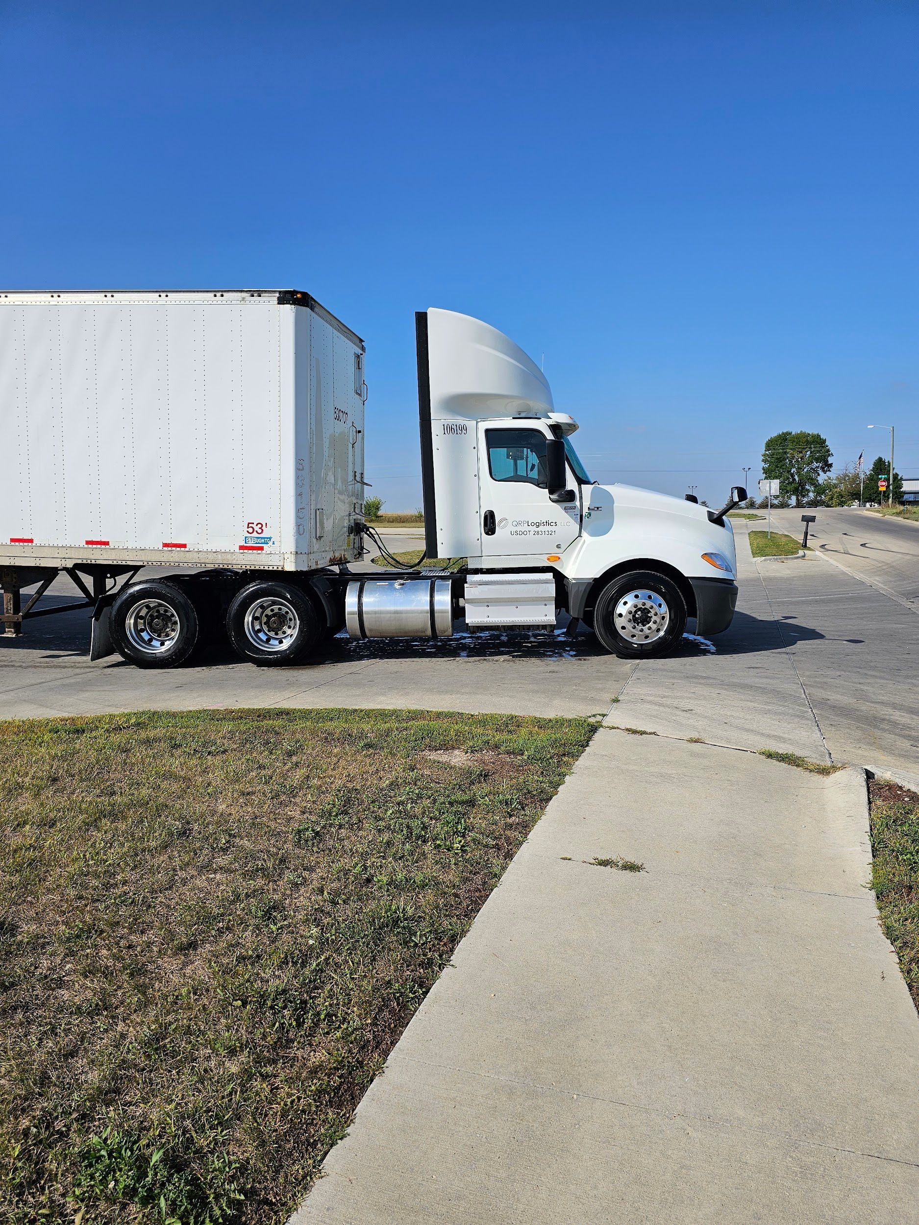 A white semi truck is parked on the side of the road next to a sidewalk.