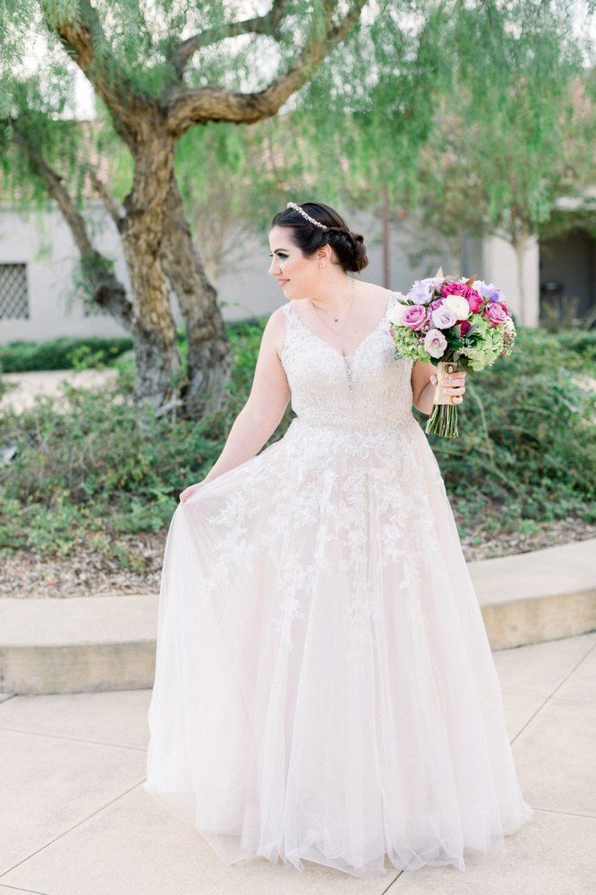 Bride in a white lace gown holding a pink bouquet outdoors by a tree
