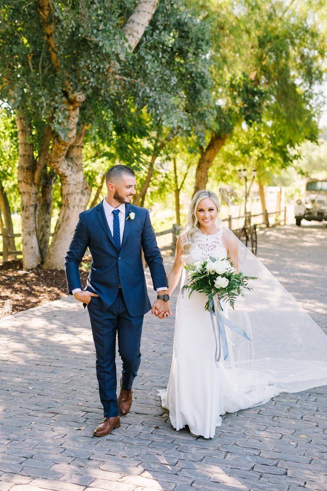 Bride and groom walking hand in hand outdoors, bride holding a bouquet, under trees on a sunny path