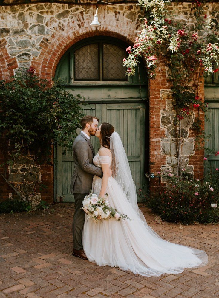 Bride and groom kiss beneath an ivy-covered brick arch with a green door and flowers.
