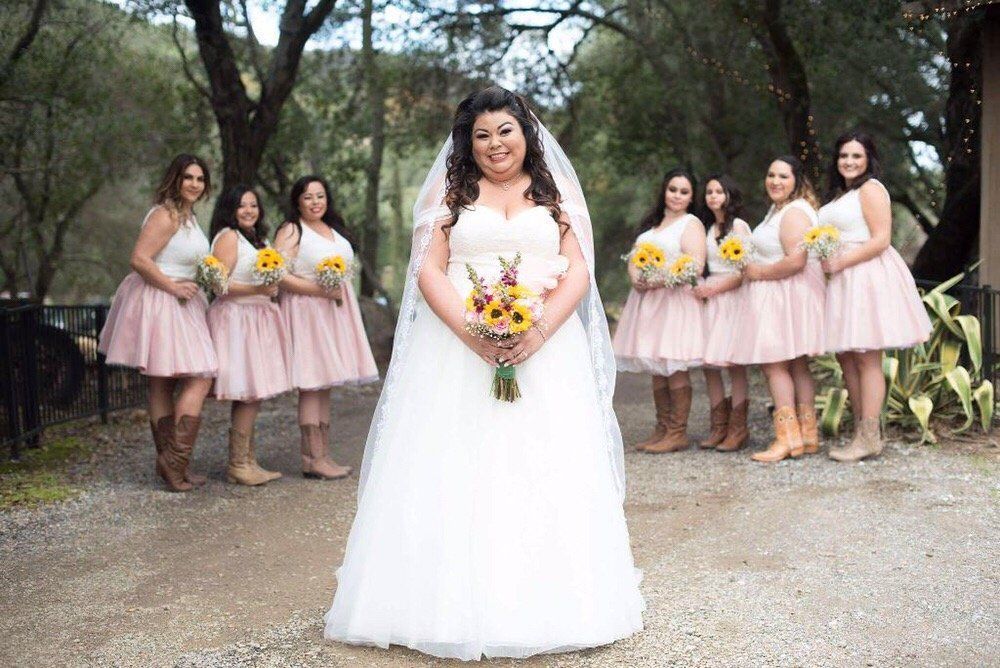 Bride in a white gown stands with bridesmaids in pink dresses holding yellow bouquets outdoors.