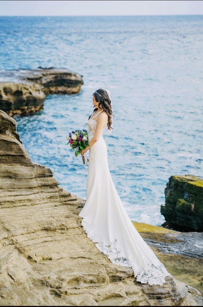 Bride in a white gown holding a bouquet on seaside rocks, facing the ocean