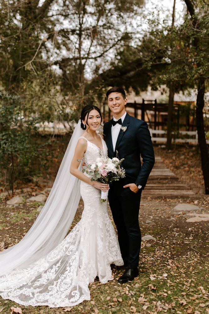 Bride and groom in wedding attire posing outdoors under trees, bride holding bouquet and veil flowing behind.