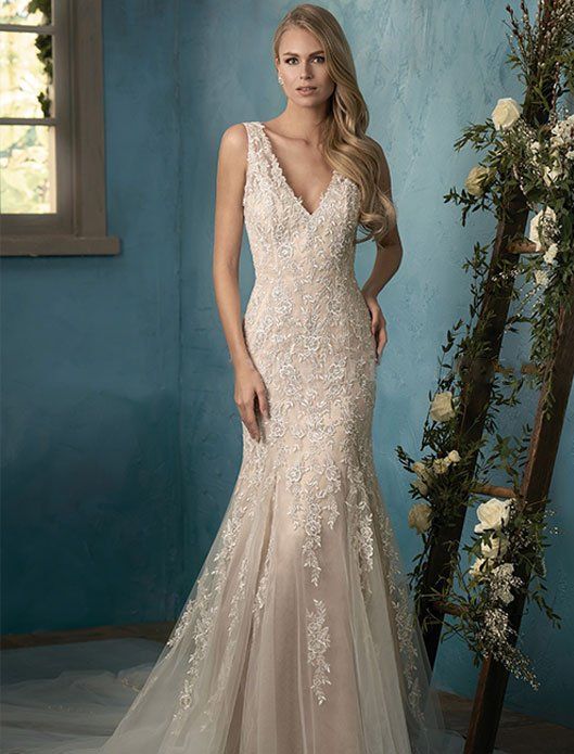 Woman in a sleeveless ivory beaded mermaid gown standing beside floral decor against a blue wall.