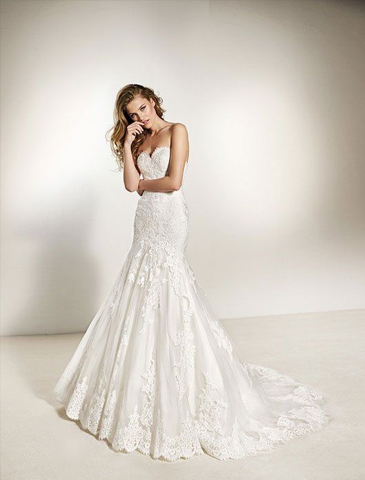 Woman in a white lace wedding dress standing against a soft beige backdrop, posing with one hand by her face.