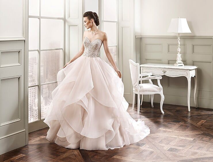 Woman in a blush ball gown standing in a bright room with a white desk and chair.