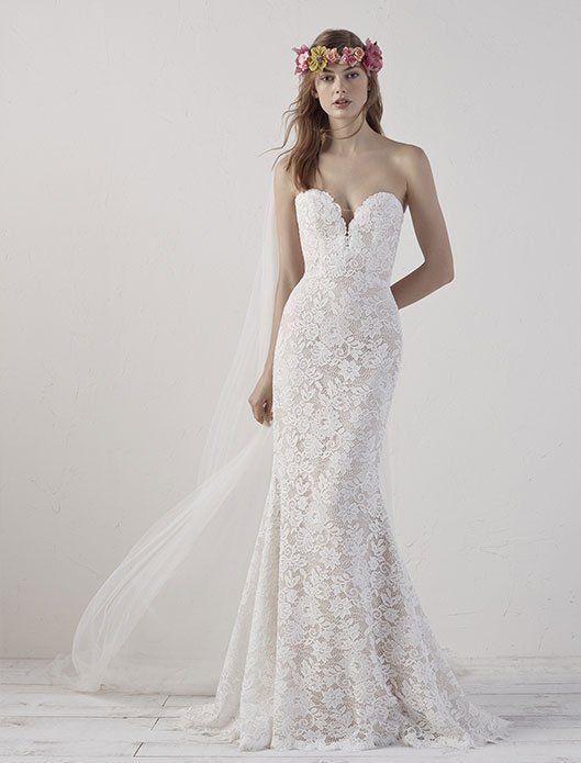 Bride in a white lace mermaid gown with a floral crown and long veil, standing against a light backdrop