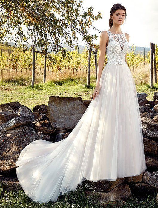 Woman in a white lace gown with a long train standing outdoors beside rocks and vineyard rows