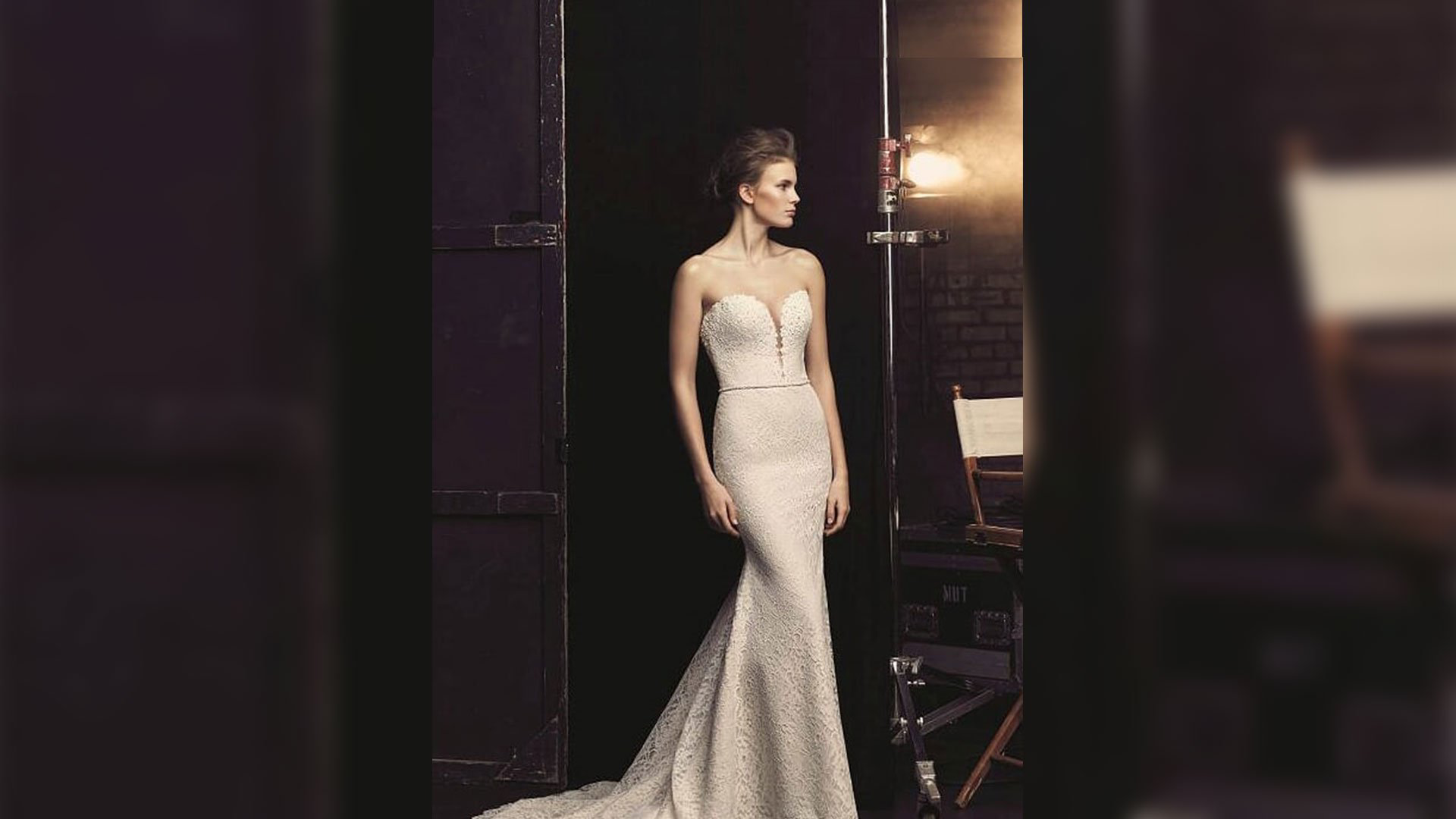 Woman in a white strapless gown standing in a dim backstage hallway by a lit mirror