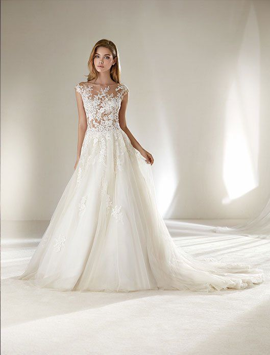 Bride in a white lace wedding gown with a long tulle train, standing in a bright studio.