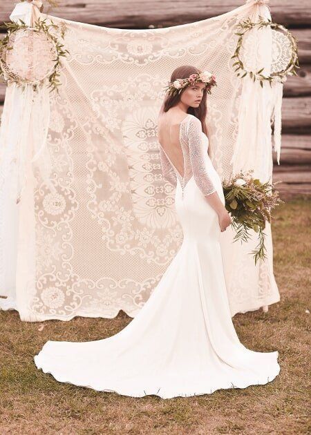 Bride in a white lace gown with long train standing before a floral backdrop outdoors