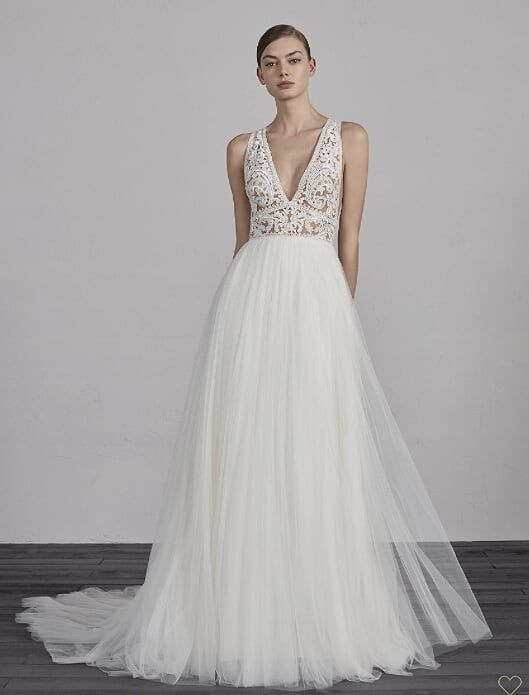 Woman in a white beaded V-neck tulle gown with a long train, standing against a gray backdrop.
