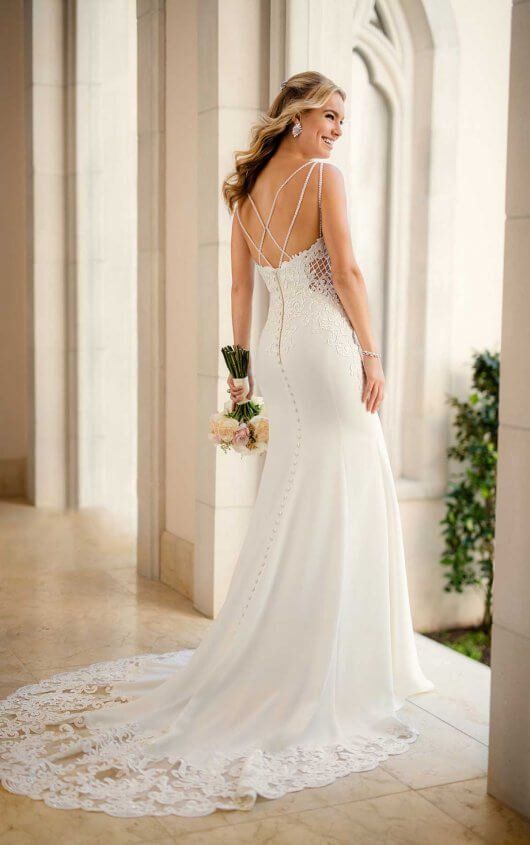 Bride in a white lace wedding gown walking in a sunlit hallway, holding a small bouquet.