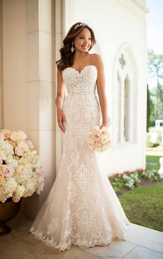 Bride in a lace white wedding dress holding a bouquet, standing outdoors by a column and flowers.