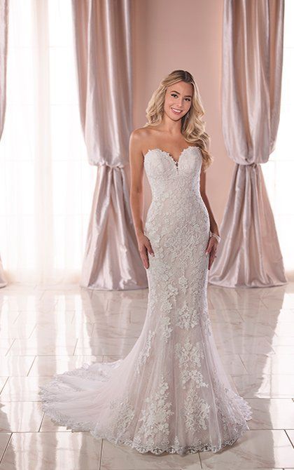 Woman in a strapless white lace wedding gown standing before pale curtains.