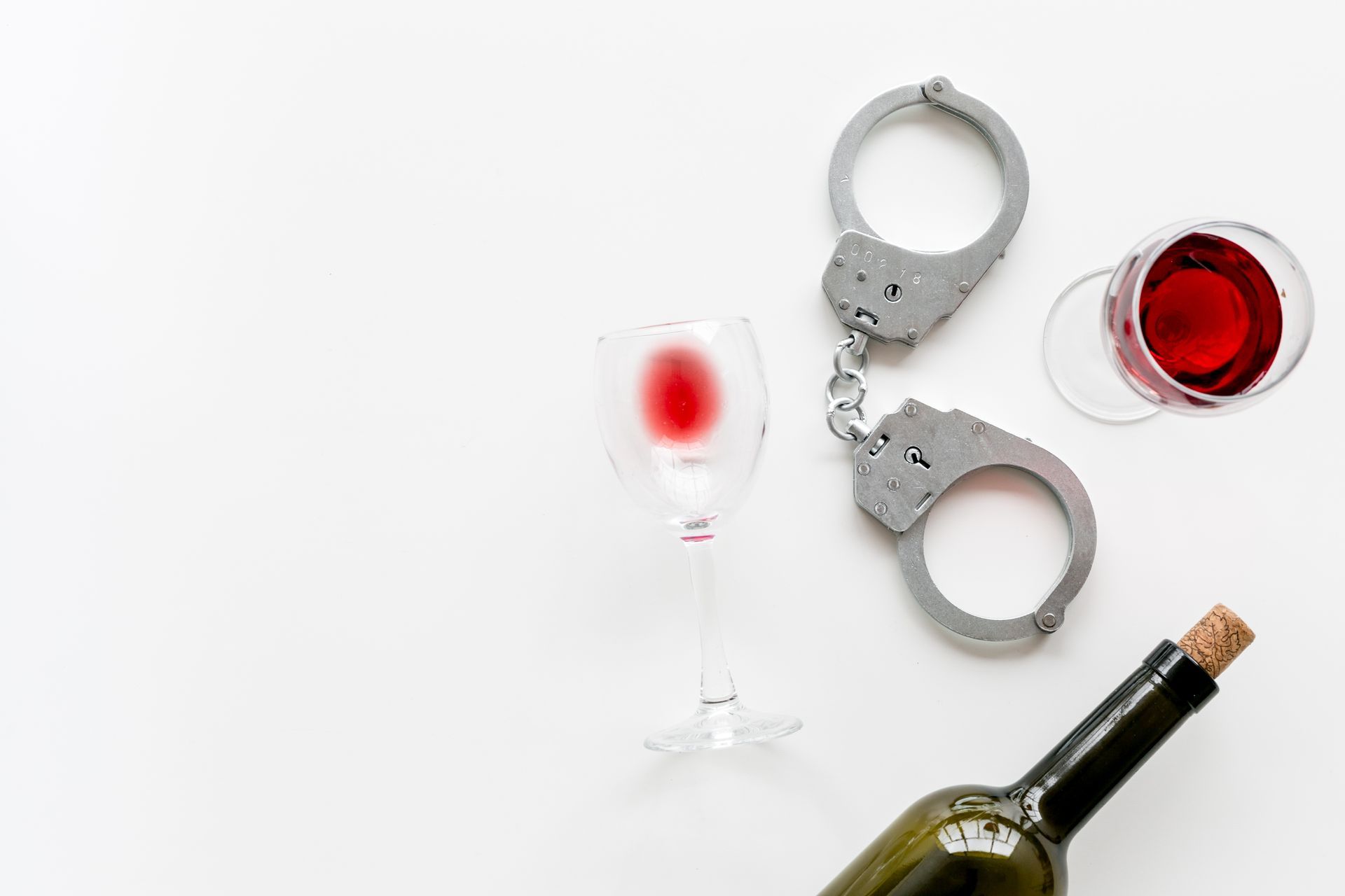 Handcuffs next to a wine bottle and two glasses of red wine, on a white surface.