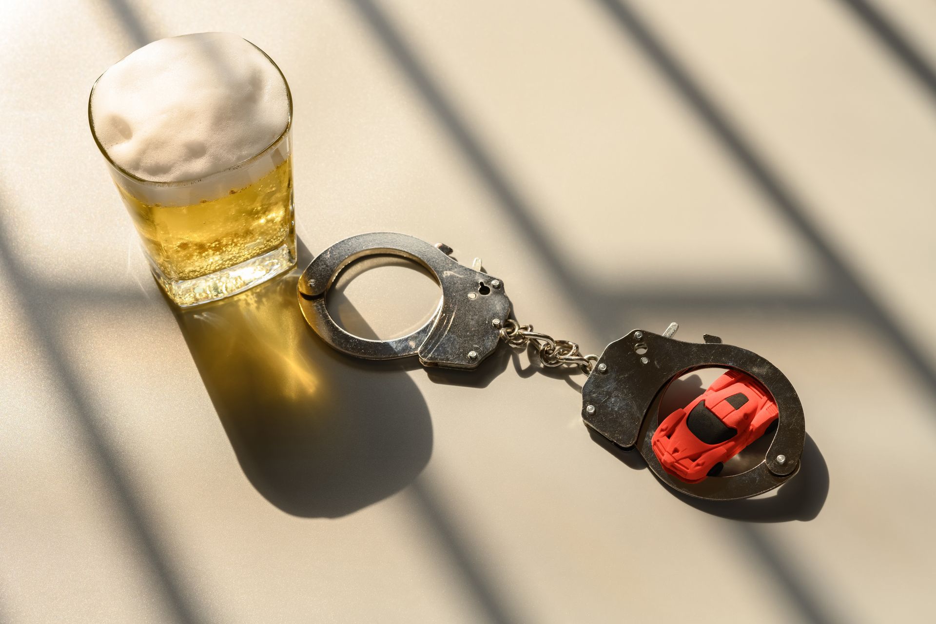 Glass of beer, handcuffs, and toy car on a surface with shadows, suggesting drunk driving.