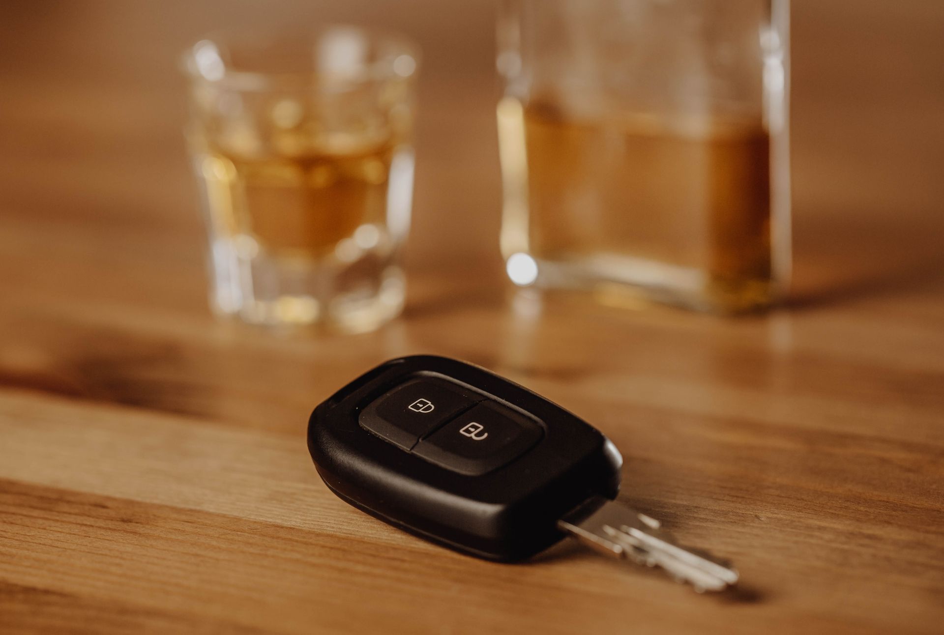 Car keys on a wooden table, with a glass and bottle of amber liquid in the background.
