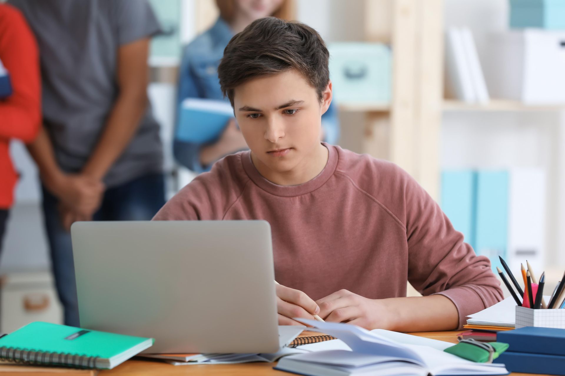 Young person focused on a laptop at a desk with notebooks and papers; other people in the background.