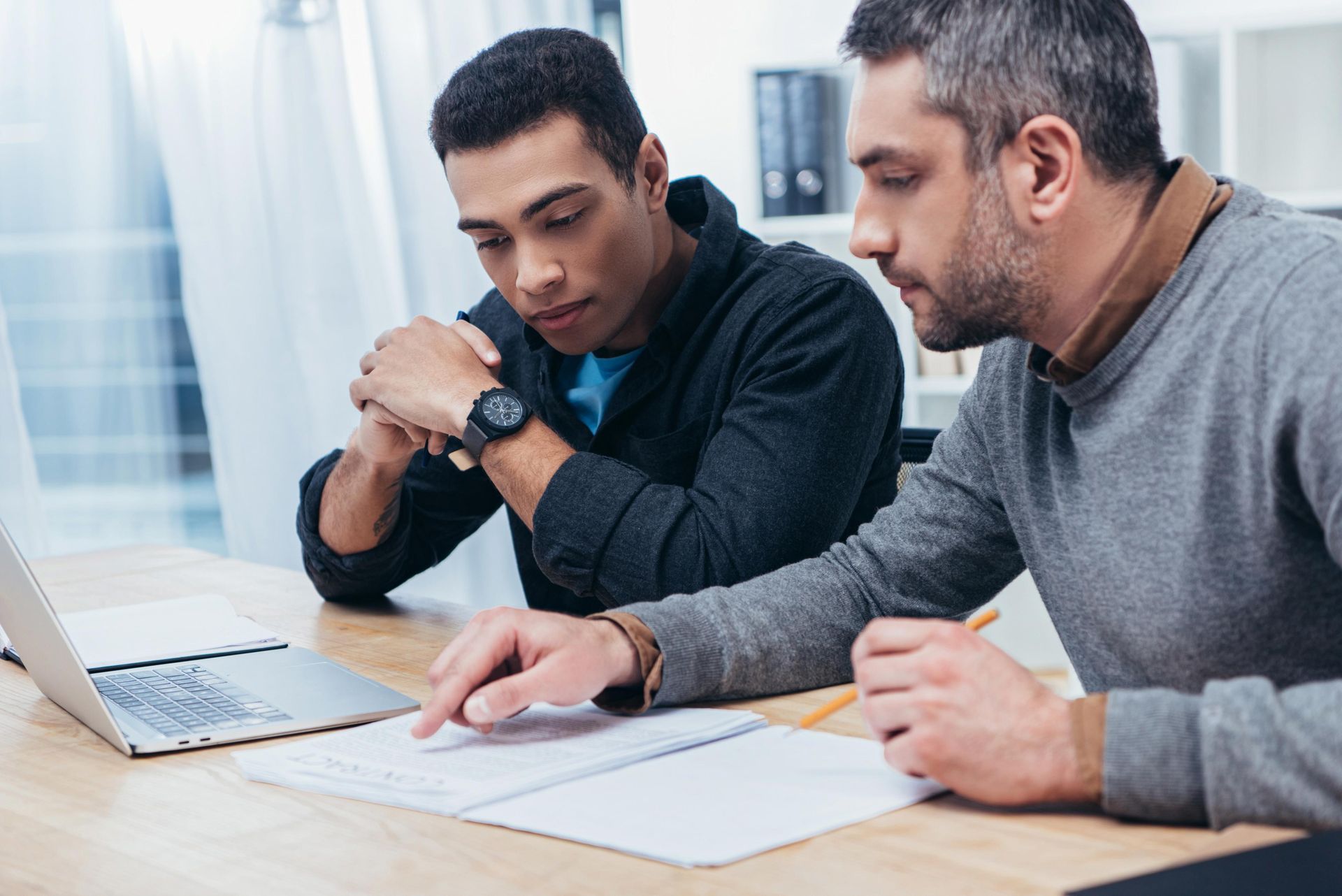 Two men at a desk, looking at a notebook and laptop. One points at the notebook.