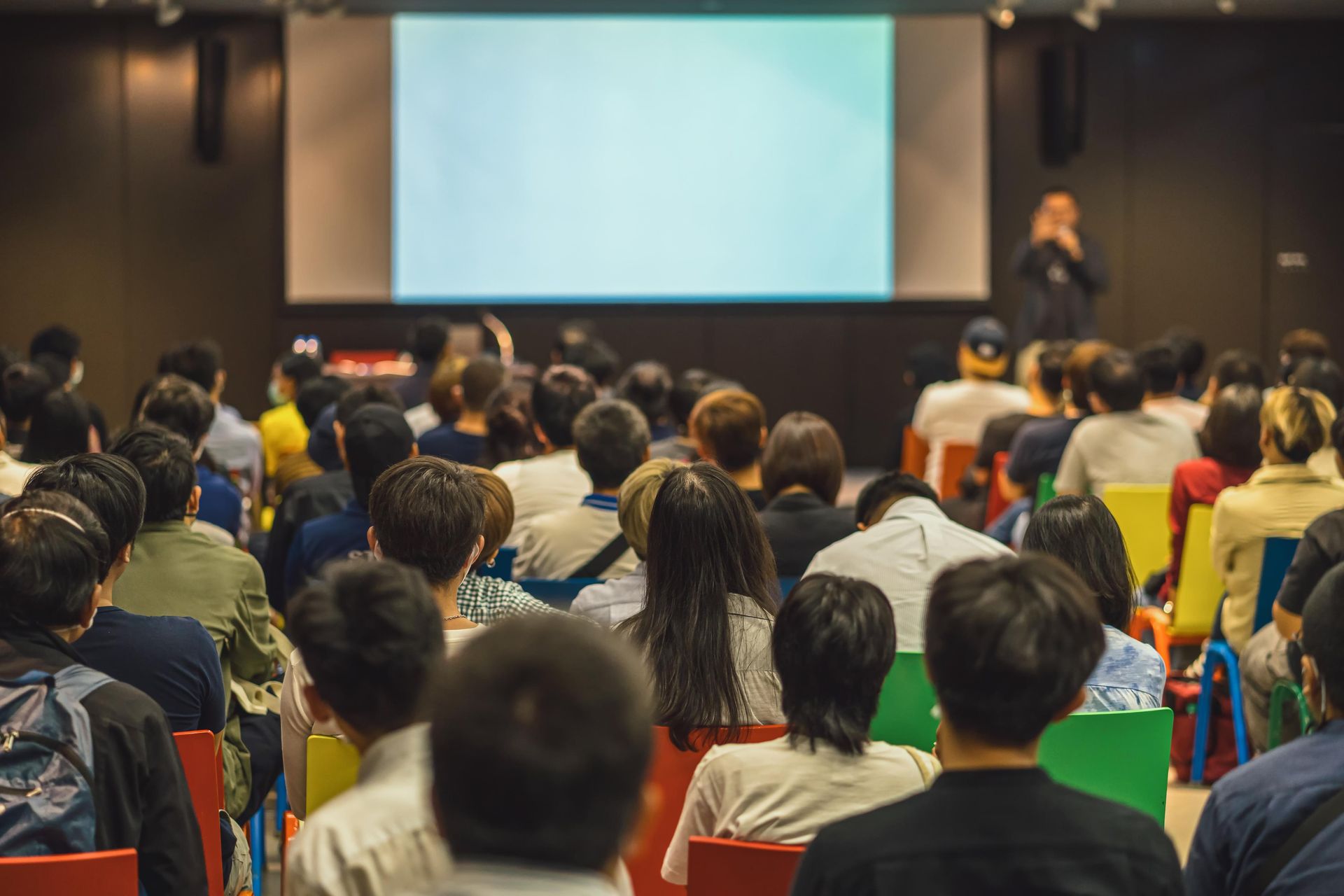 Audience seated in a lecture hall facing a blank screen; speaker in background.