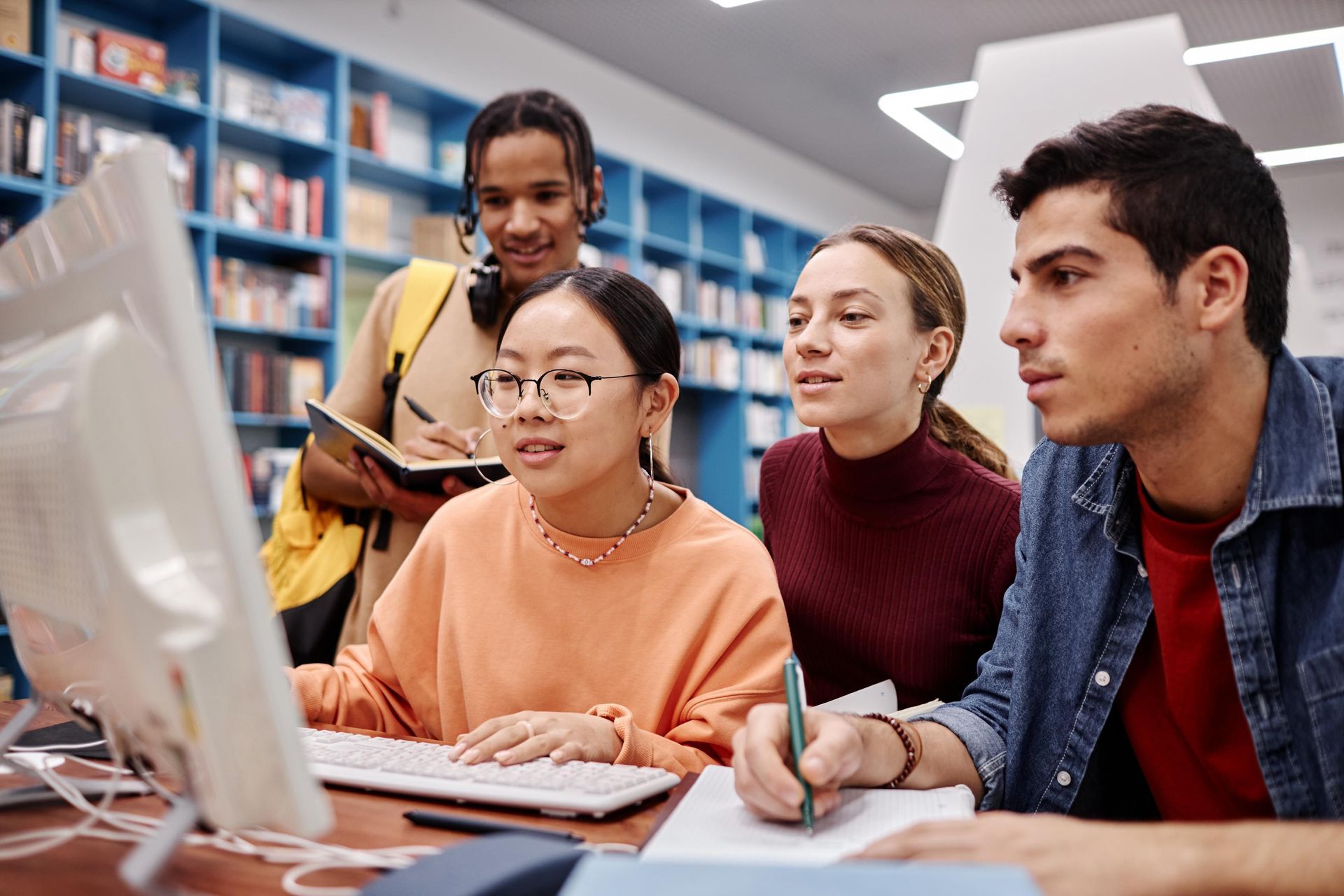 Students collaborating around a computer in a library. One types, three look on, one holds a notepad.