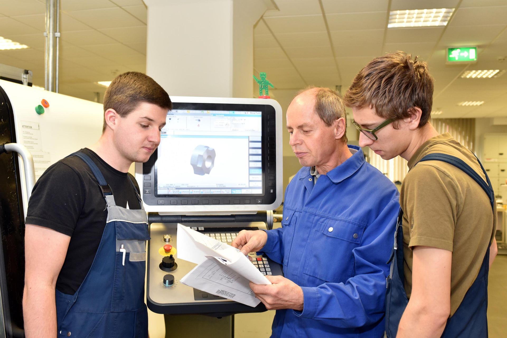 Three people looking at plans near a machine; one points at a computer screen showing a 3D image.