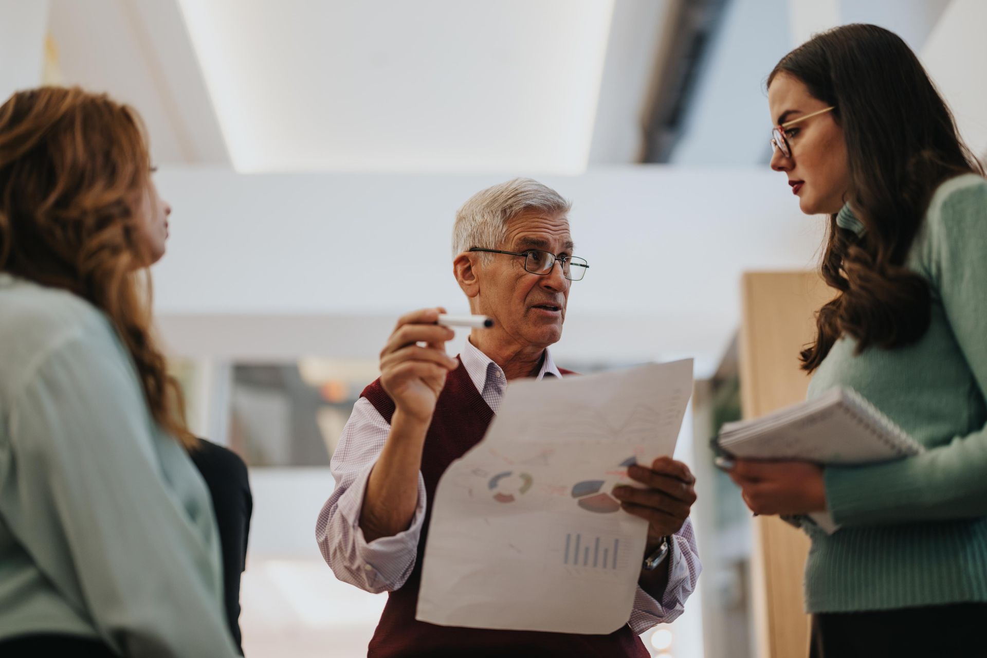Three people in office setting; man gestures, holds paper. Two women listen intently.