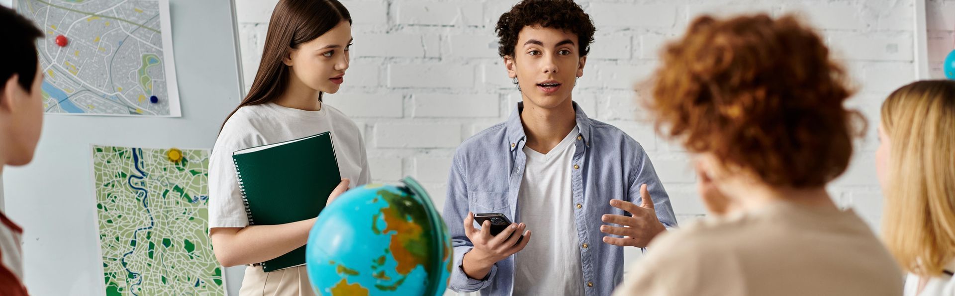 Students in a classroom. A young man speaks while holding a phone and gesturing. There's a globe and maps on the wall.