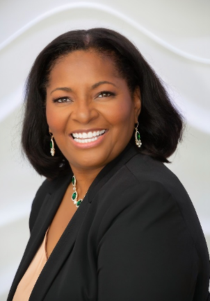 Woman with dark hair smiles, wearing a black blazer, green jewelry, against a white background.