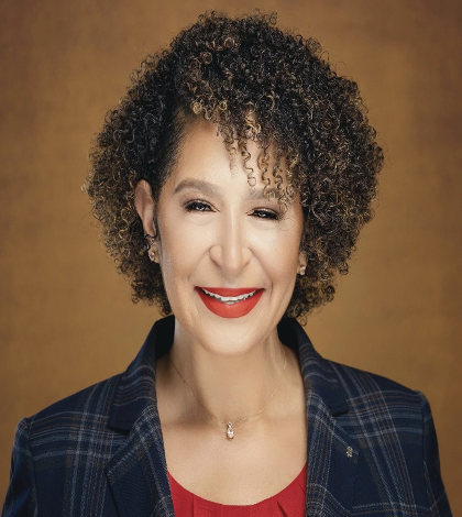 Woman with curly brown hair, wearing a blazer over a red top, smiling with red lipstick.