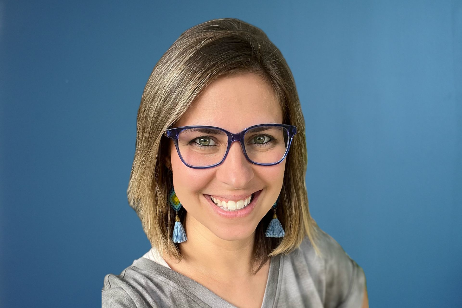 A woman wearing glasses and earrings is smiling in front of a blue background.