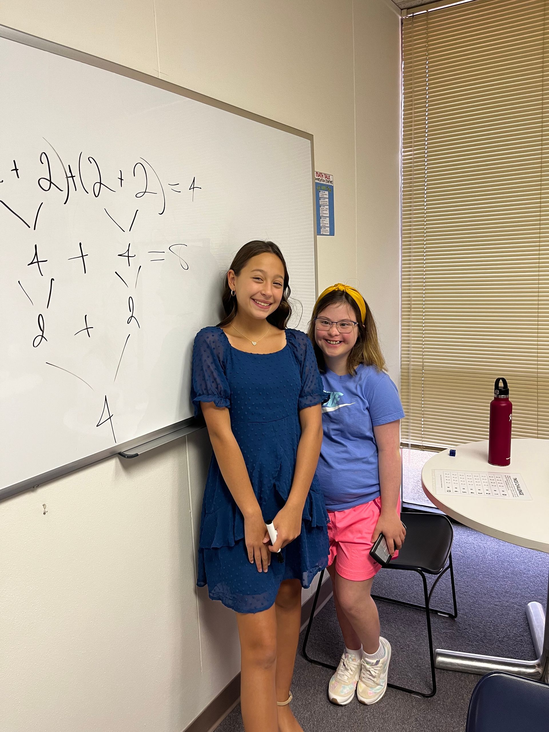 Two young girls are standing next to each other in front of a whiteboard.