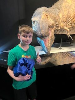 A young boy is standing next to a dinosaur statue and giving a thumbs up.