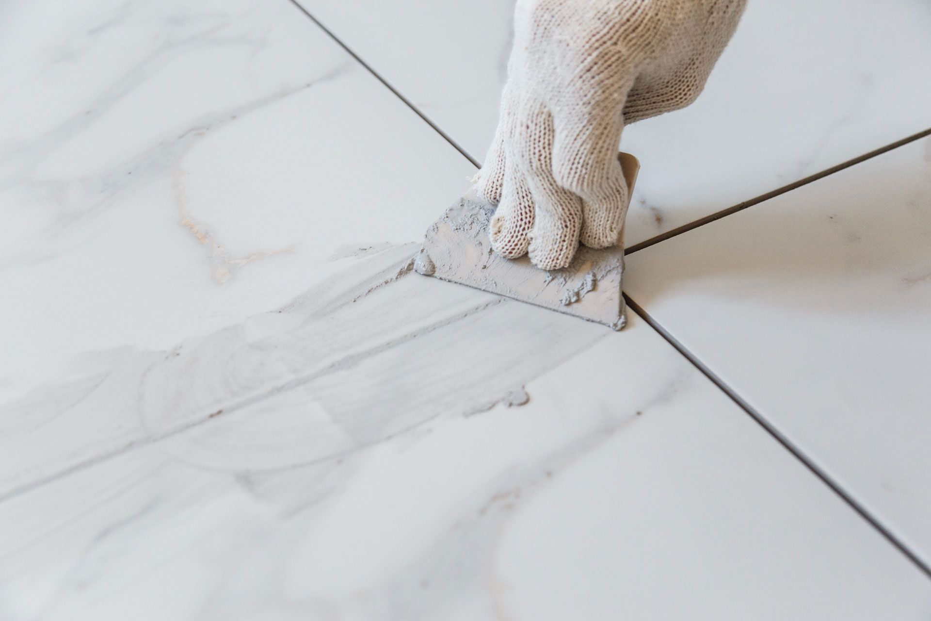 Gloved hand using a trowel to apply grout between white marble tiles.