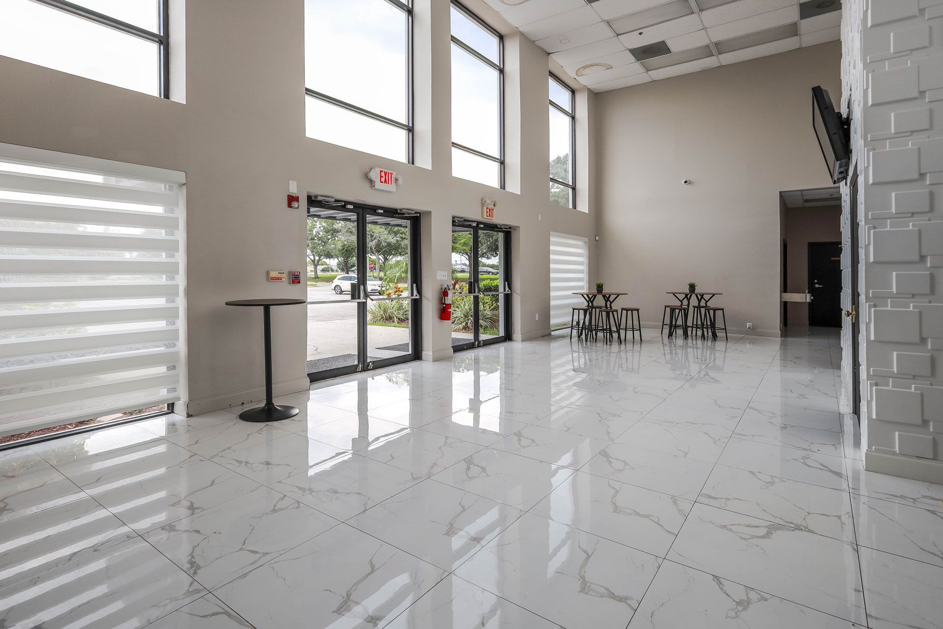 Lobby with white tile floor, tall windows, and a black door leading outside. A small table and chairs are in the background.