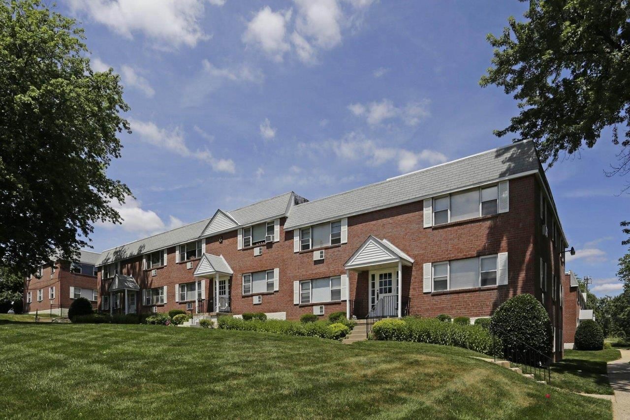 Exterior view of a brick apartment building with green lawn and trees surrounding it