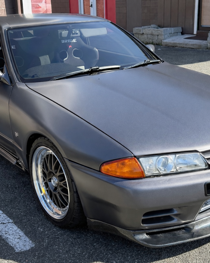 A matte gray Nissan Skyline R32 GT-R with aftermarket multi-spoke wheels parked on an asphalt lot.